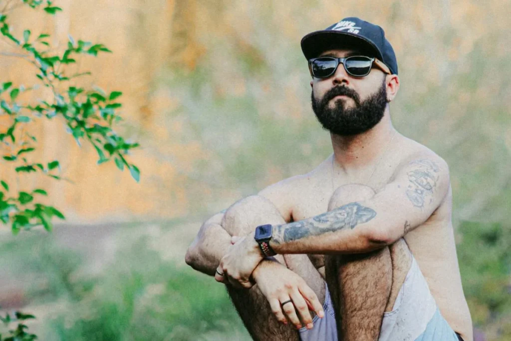 A man sits with sunglasses, beard, and cap